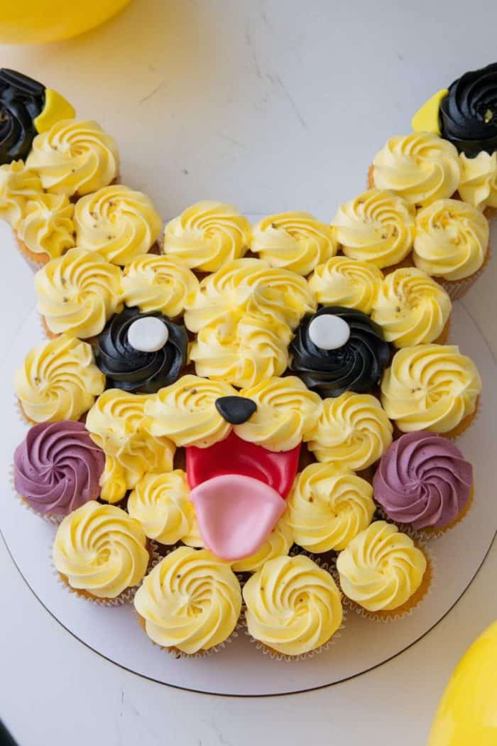 Cupcakes arranged and frosted to resemble a popular cartoon character with yellow icing, black and white eyes, pink cheeks, and a red tongue. The design forms a playful face with large ears.