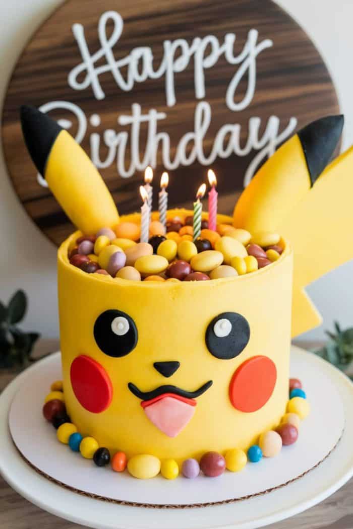 A Pikachu-themed birthday cake adorned with three lit candles on top. The cake is decorated with Pikachu's face and ears, and surrounded by colorful candies. A "Happy Birthday" sign is in the background.