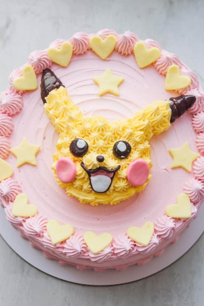 A round cake with pink icing, decorated with yellow star and heart shapes. The center features an icing design of a character's face with yellow ears, large eyes, and a wide smile, resembling Pikachu.