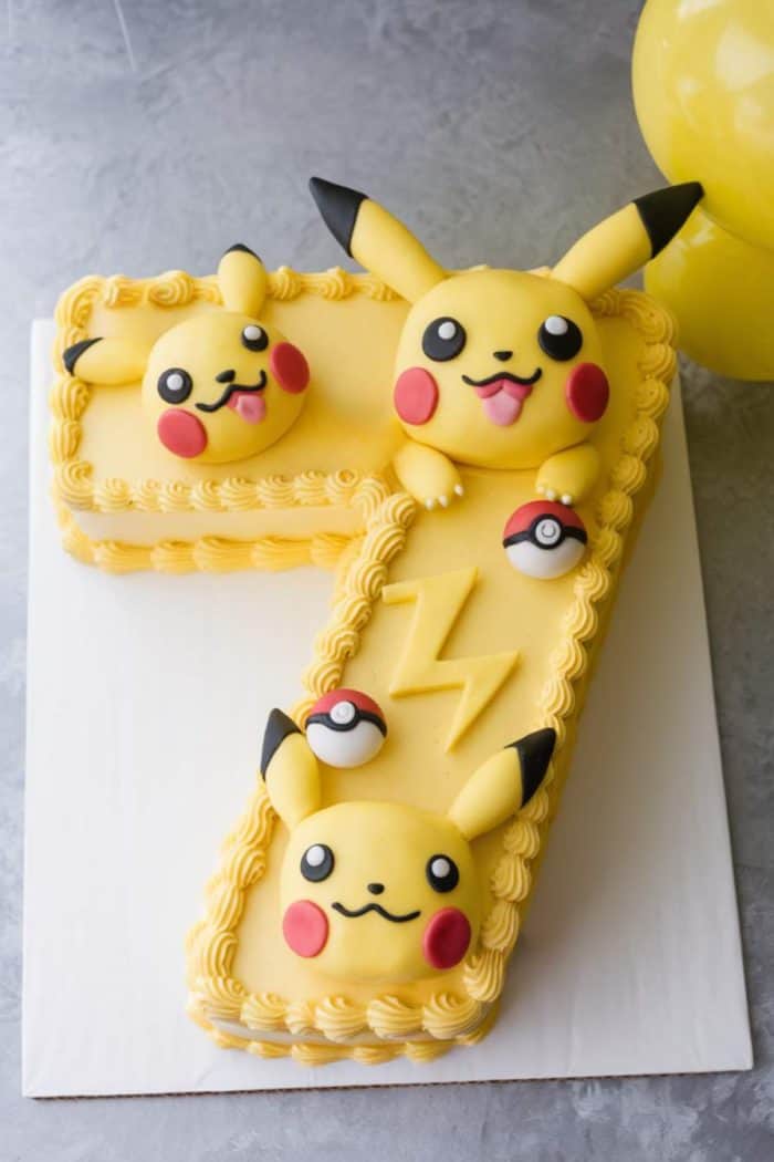 A cake shaped like the number seven, decorated with yellow icing and featuring Pikachu faces and tiny Poké Balls. The cake's surface has a zigzag lightning pattern, and it is placed on a white base.