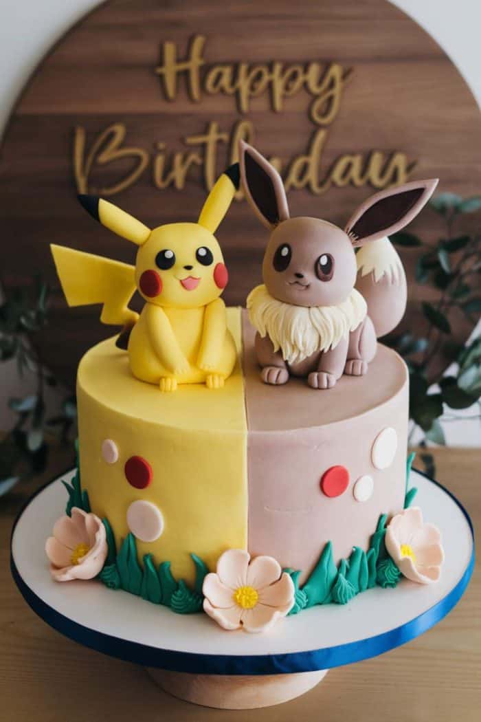 A two-toned cake featuring fondant figures of Pikachu and Eevee atop. The left side is yellow with Pikachu, and the right is brown with Eevee. Decorated with green leaves and pink flowers. "Happy Birthday" is written in the background.