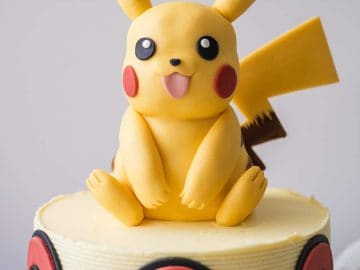 A round cake decorated with Poké Balls around the sides and topped with a Pikachu figurine made from fondant, showing Pikachu sitting with its tongue out and ears upright.
