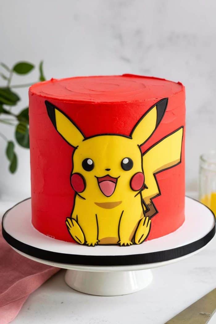 A cake with red frosting features a design of Pikachu, the yellow Pokémon character, on the side. It sits on a white platter, with a pink cloth and green plant in the background.