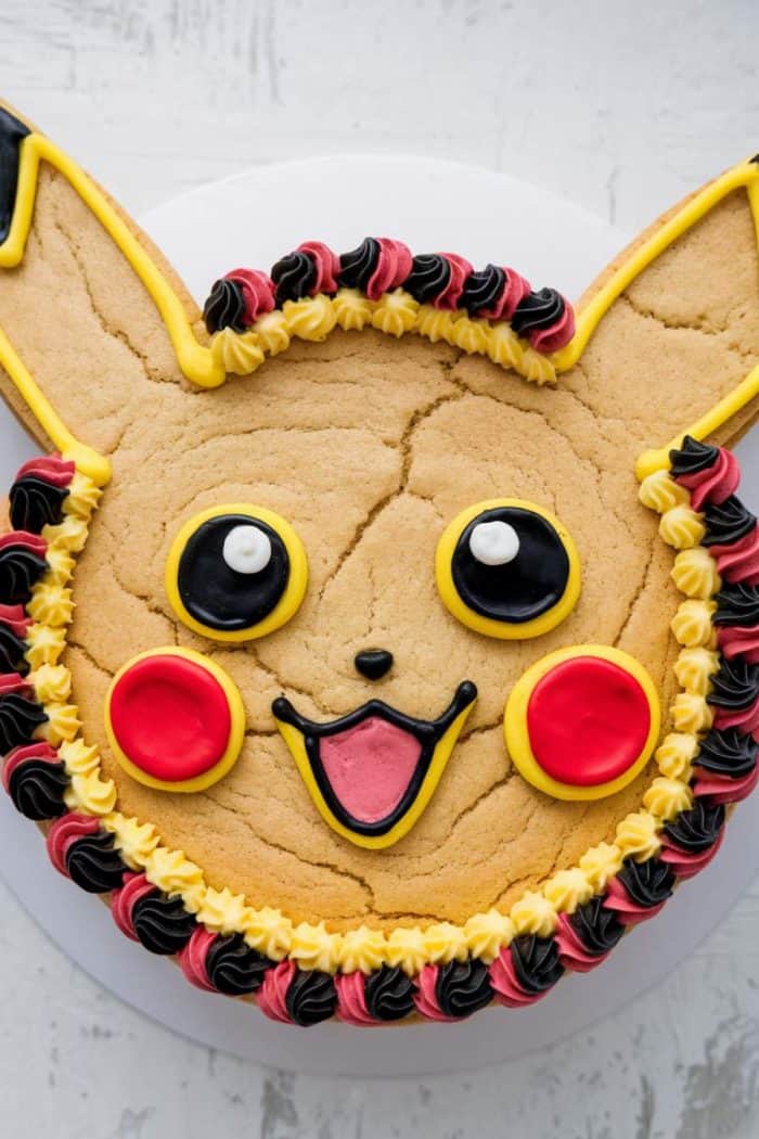 A cake decorated to resemble a cartoon character with large ears and a smiling face. It features yellow, black, pink, and red icing for details like eyes, mouth, and cheeks, with colorful frosting trim around the edges.