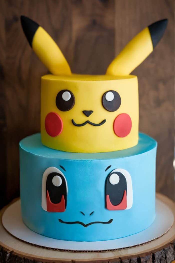 A two-tier cake decorated as cartoon characters. The top tier is yellow with ears and a face with big eyes and red cheeks. The bottom tier is blue with a smiling face and large eyes. The cake is on a wooden cake stand.