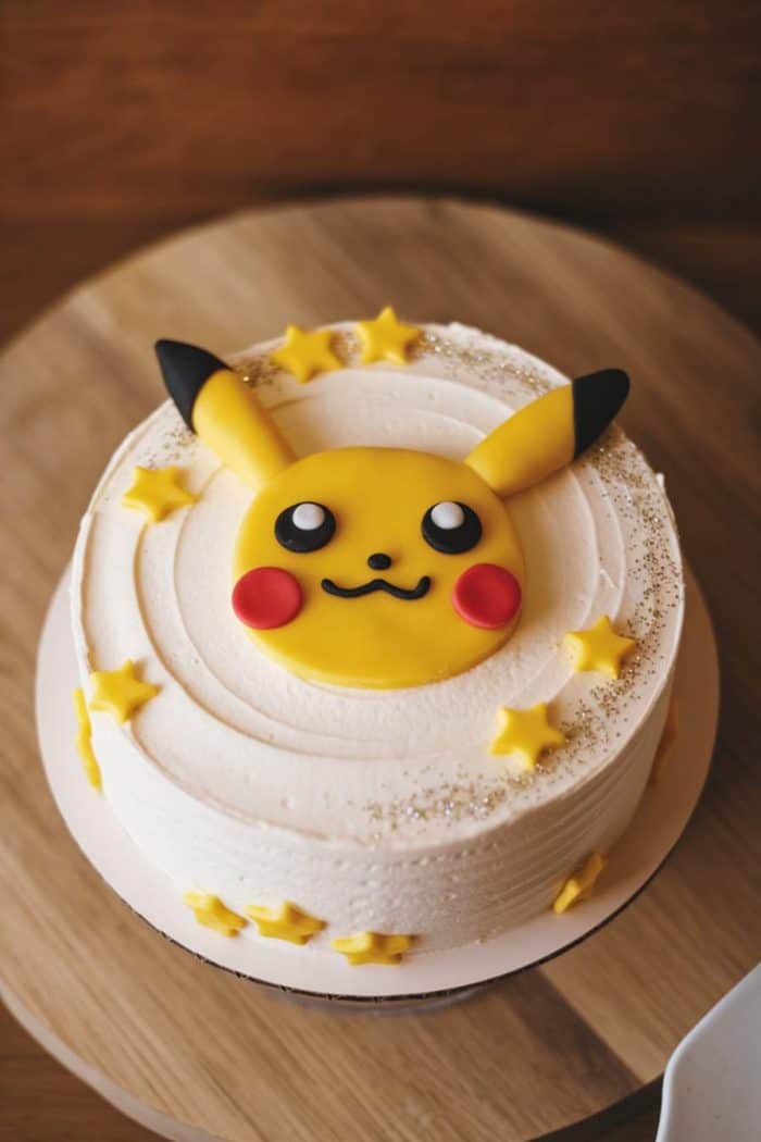 A round, white cake decorated with a fondant character face on top, featuring yellow ears and red cheeks. The cake is adorned with small yellow star shapes around its edge, placed on a wooden surface.