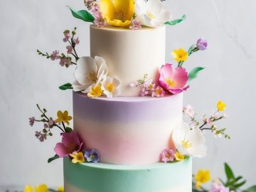 A three-tiered cake with pastel gradient layers of blue, purple, and pink. It's elegantly decorated with vibrant sugar flowers in yellow, white, and pink, displayed on a white cake stand. Surrounded by flowers and green leaves on the table.