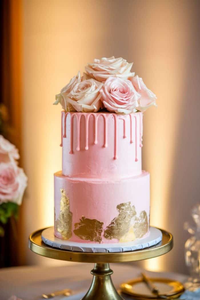 A two-tiered pink cake with elegant drip icing, adorned with gold leaf accents. The top is decorated with a bouquet of pale pink roses. The cake is displayed on a gold stand in a softly lit setting.