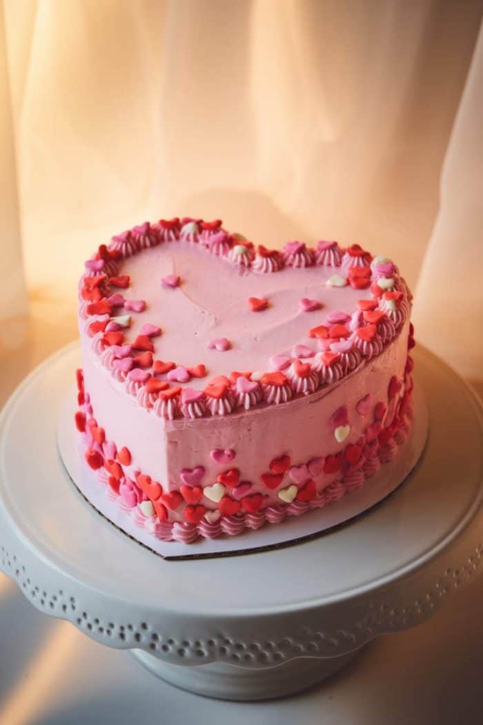 A heart-shaped cake with pink frosting is adorned with red and pink heart-shaped sprinkles, sitting on a round white cake stand. The background features soft, warm lighting.
