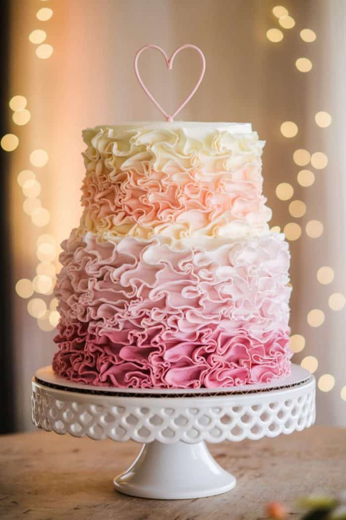 A three-tiered cake with ruffled icing in ombre shades of pink, from dark to light, sits on a decorative white cake stand. A simple heart topper adorns the cake. Soft, blurry lights twinkle in the background, creating a warm ambiance.