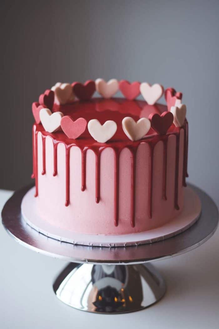 A pink cake with red drip icing on a silver stand, decorated with a circle of heart-shaped toppers in red, pink, and white.
