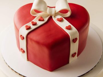 Heart-shaped cake covered in red fondant with a white fondant ribbon and bow. Small red heart decorations line the ribbon.