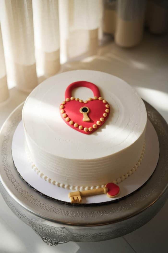 A round white cake on a decorative silver stand, adorned with a red heart-shaped lock design on top. A matching red key lies beside it, adding a whimsical touch. Sunlight softly illuminates the scene.