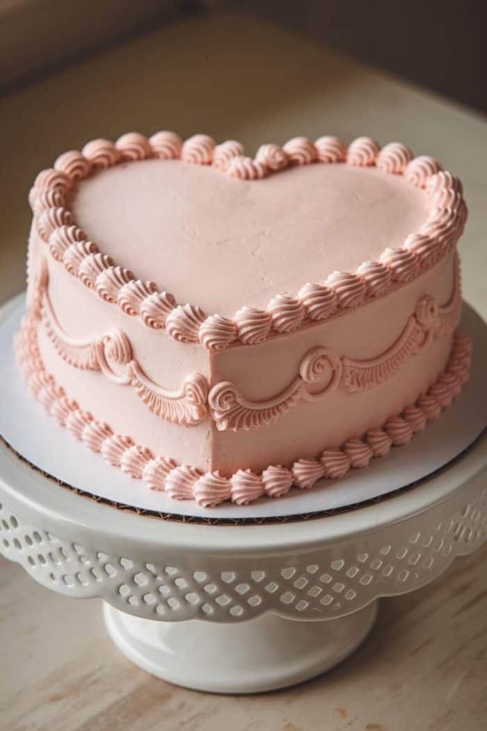 Heart-shaped cake with light pink frosting, decorated with scalloped and piped details on the sides and edges, placed on a white, lattice-edged cake stand.