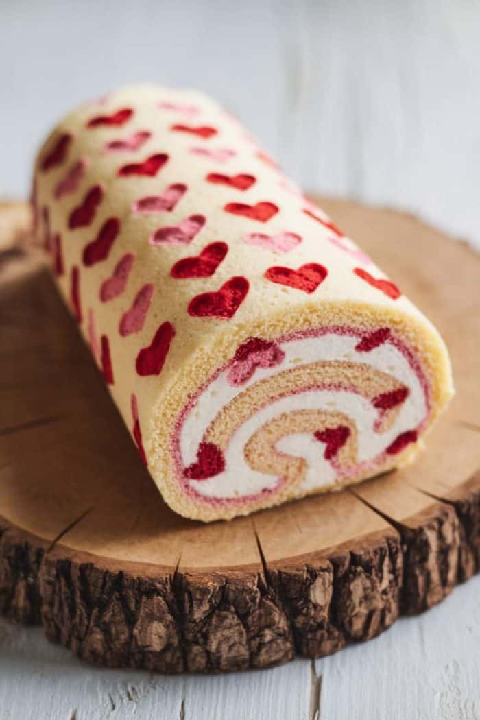 A decorative Swiss roll cake with a light cream filling and raspberry swirls sits on a wooden cake board. The cake's exterior is patterned with red heart shapes.