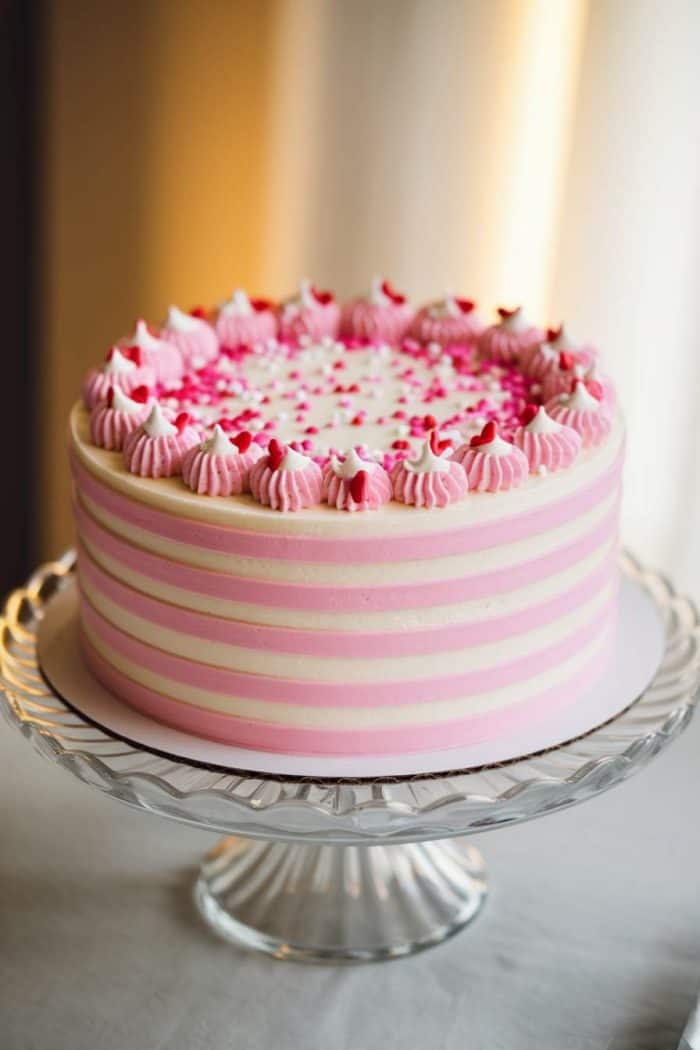 A cake with pink and white striped frosting, topped with pink whipped dollops and scattered pink heart sprinkles, sits on a glass stand.