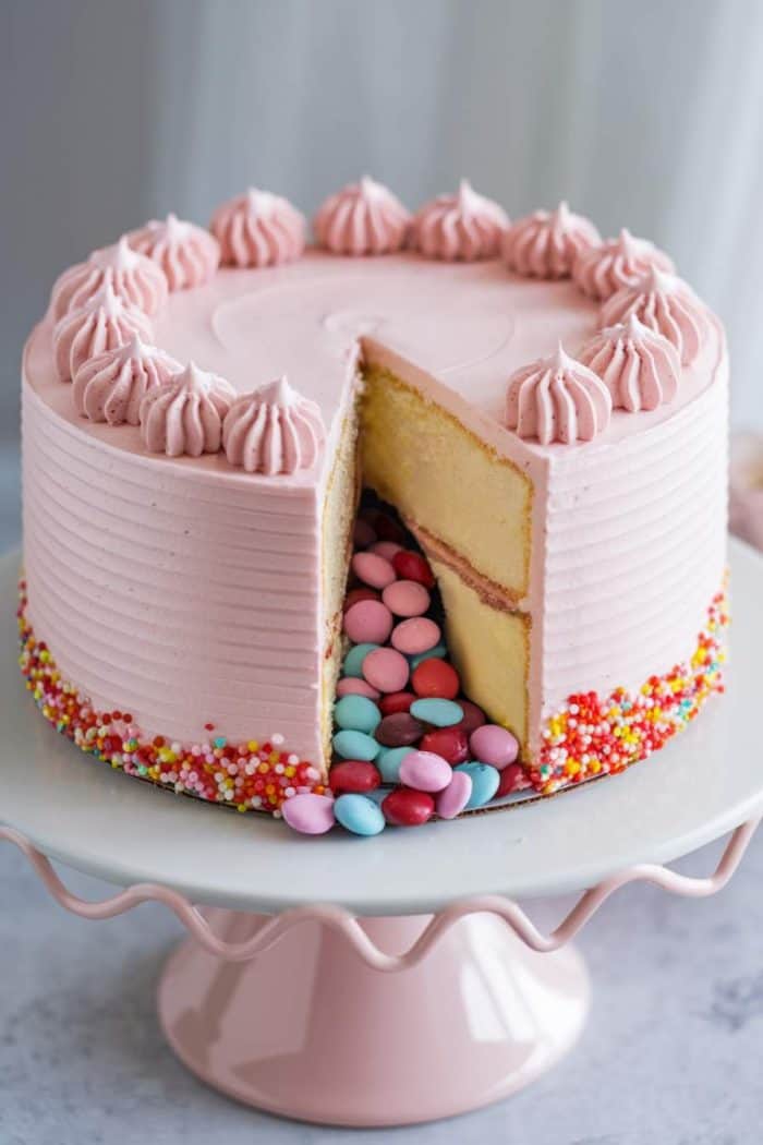 A pink frosted cake with a slice removed, revealing colorful candy inside. The cake has piped frosting swirls on top and is decorated with multicolored sprinkles around the base, sitting on a white, wavy-edged cake stand.