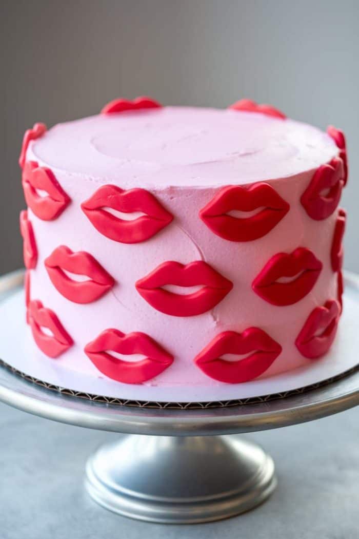 A pink frosted cake decorated with red and pink fondant lips is displayed on a silver cake stand. The cake has a smooth finish, and the lips are evenly spaced around the sides.
