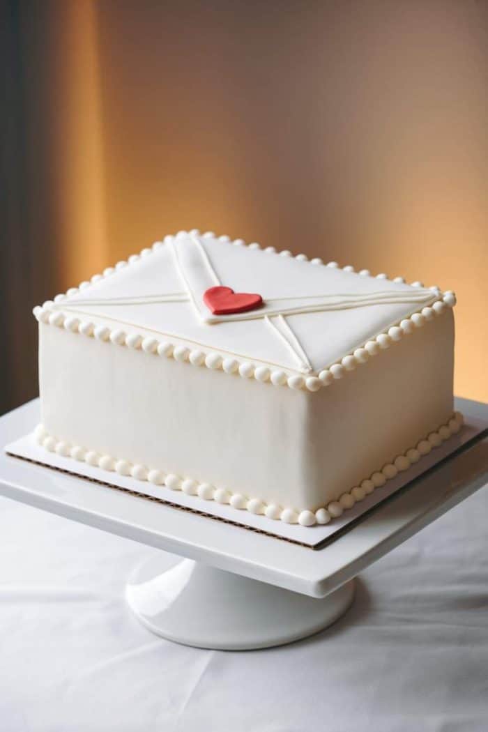 A rectangular cake designed to look like an envelope, covered in white fondant with a pearl border. A small red heart is placed in the center. The cake is displayed on a white stand against a softly lit background.