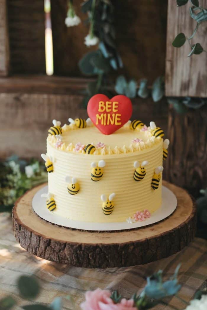 A yellow frosted cake with bee decorations is displayed on a wooden slab. A red heart-shaped topper with the words "BEE MINE" is placed on top, surrounded by small decorative bees and flowers.