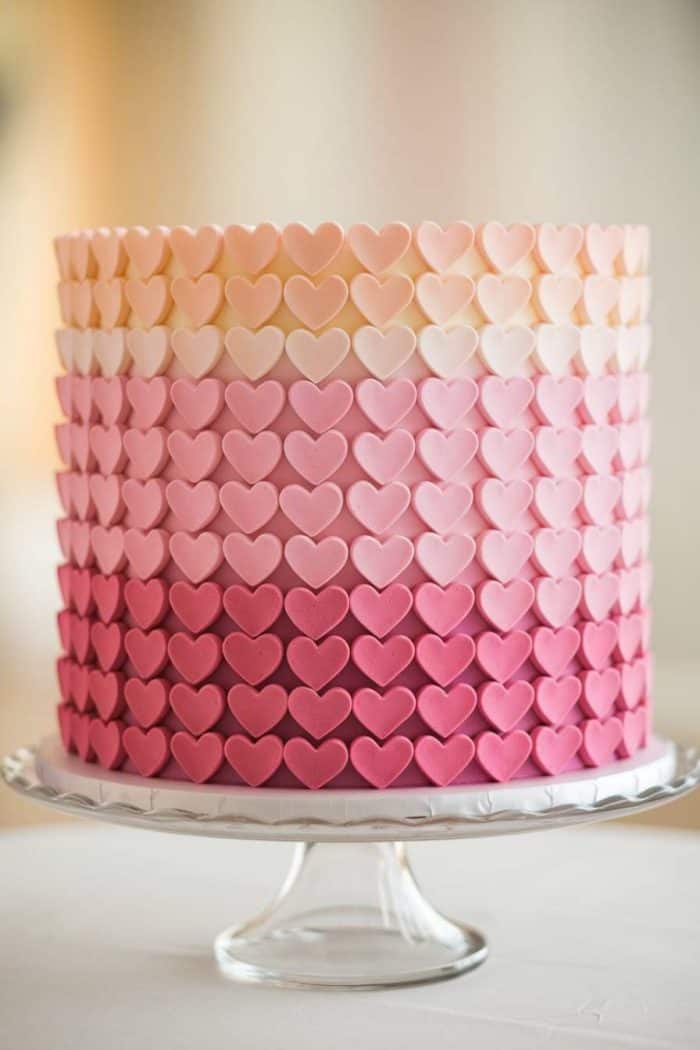 A tall cake adorned with rows of heart-shaped fondant pieces in a gradient pattern, transitioning from light cream at the top to deep pink at the bottom. The cake is displayed on a clear glass stand against a soft, neutral background.