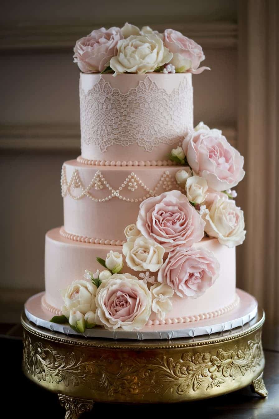 A three-tiered pink wedding cake adorned with intricate lace designs and decorated with large, delicate pink and white roses. The cake is displayed on an ornate gold stand.