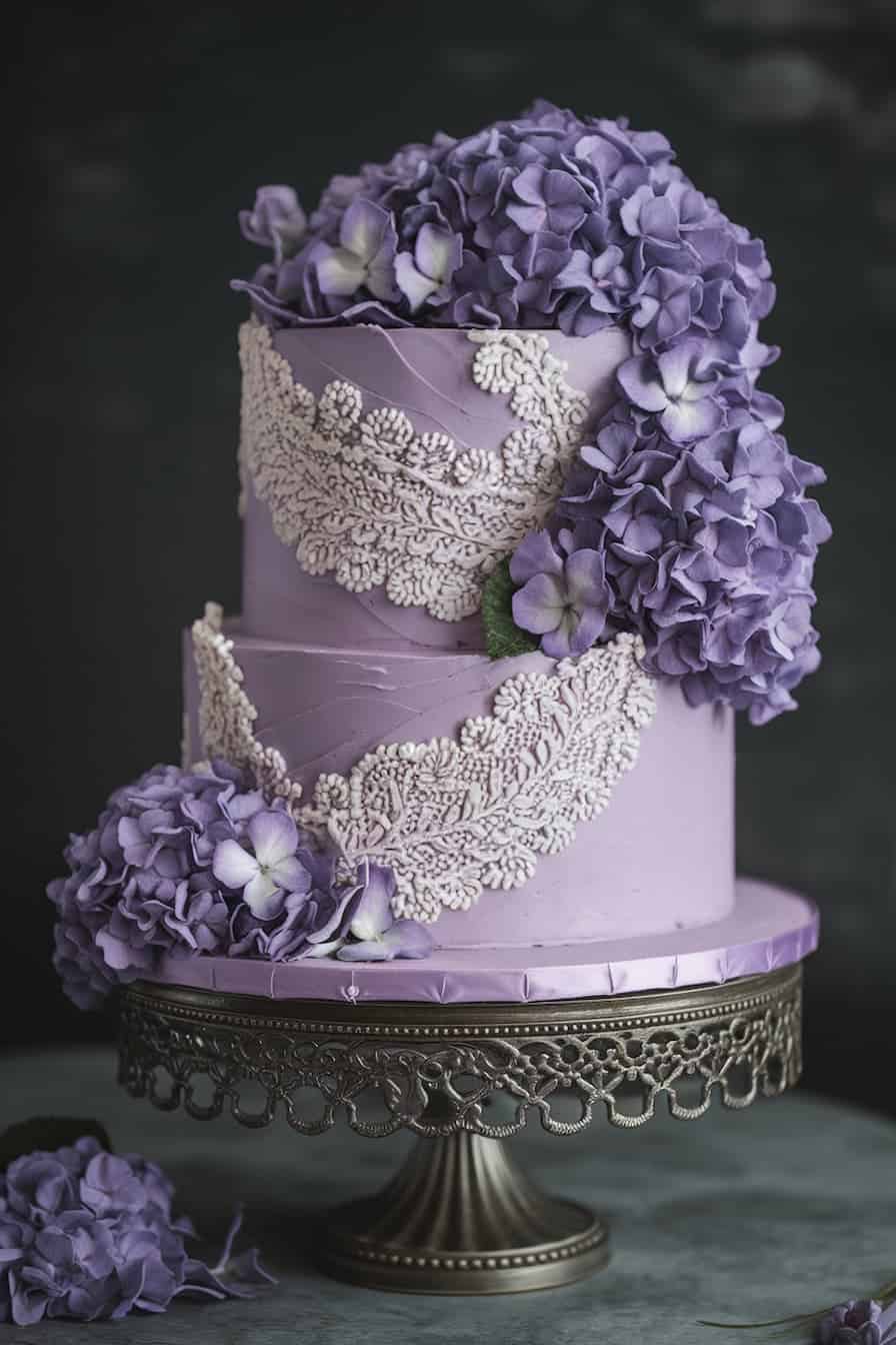 A two-tiered purple cake adorned with intricate white lace-like icing and cascading purple hydrangeas, placed on an ornate metal cake stand. The background is dark, highlighting the cake's elegant design.
