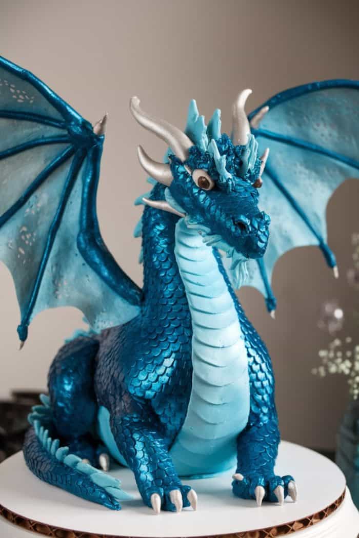 A detailed, realistic cake designed to look like a blue dragon with silver horns and wings. The dragon sits upright on a white base, displaying intricate scales, fierce eyes, and an expressive face.