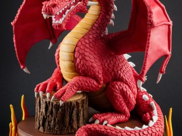 A detailed cake featuring a red dragon with white horns and claws, sitting atop a brown base. The dragon has extended wings and fierce yellow eyes. The cake's base is decorated with red and orange flames.