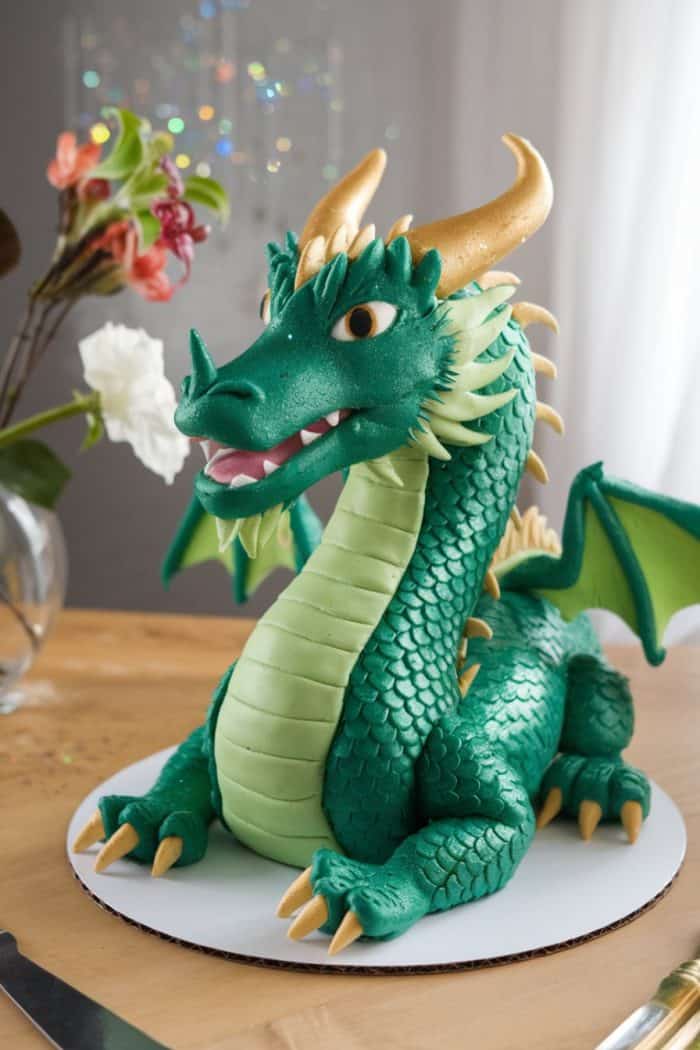 A detailed, sculpted cake resembling a green dragon with golden horns and wings sits on a wooden table. The dragon has a textured, scaly body and bright eyes. A vase with flowers is visible in the background.