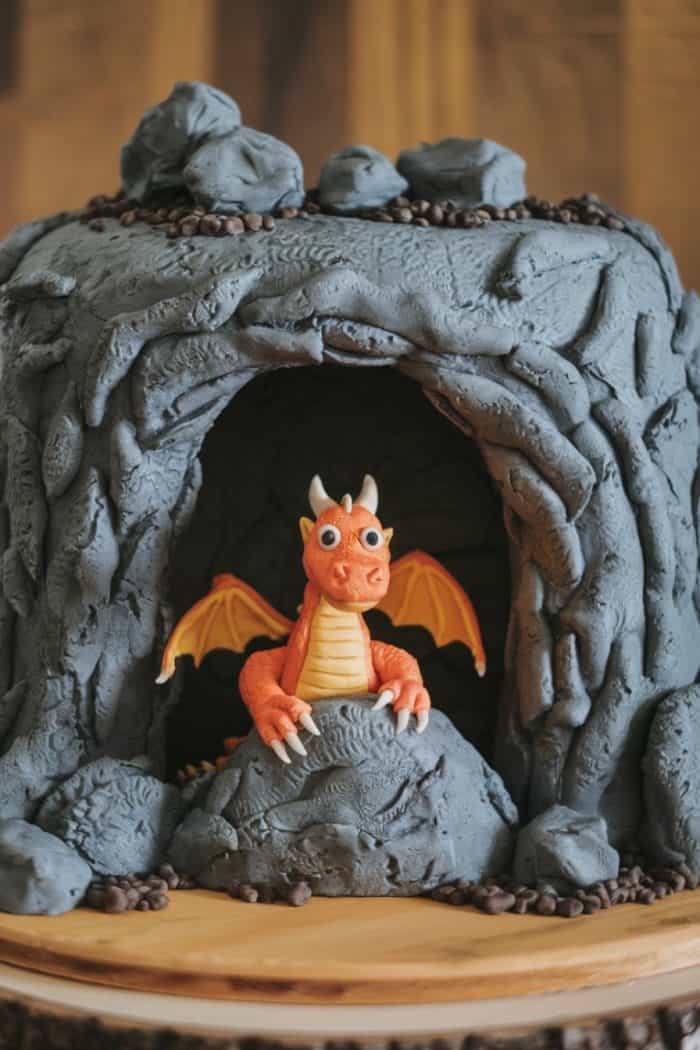 A cake designed as a rocky cave with a small, cute dragon figurine peeking out. The dragon is orange with yellow wings, and the cave is textured gray. The cake sits on a wooden surface.