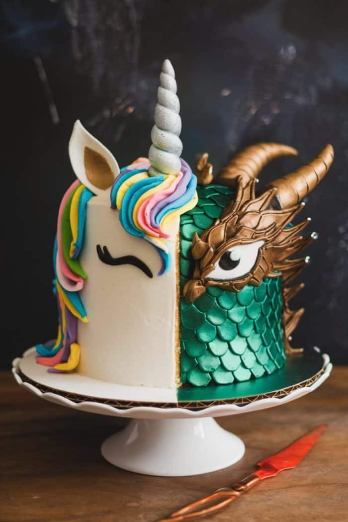 A whimsical cake with a unicorn design on the left half, featuring a pastel-colored mane and a silver horn, and a dragon theme on the right half, displaying green scales and brown horns. It sits on a white cake stand.