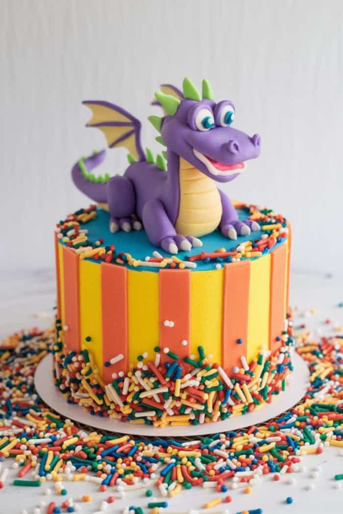 A colorful cake topped with a purple dragon figurine. The cake has vertical orange and yellow stripes and is decorated with rainbow sprinkles on top and scattered around the base on a white surface.