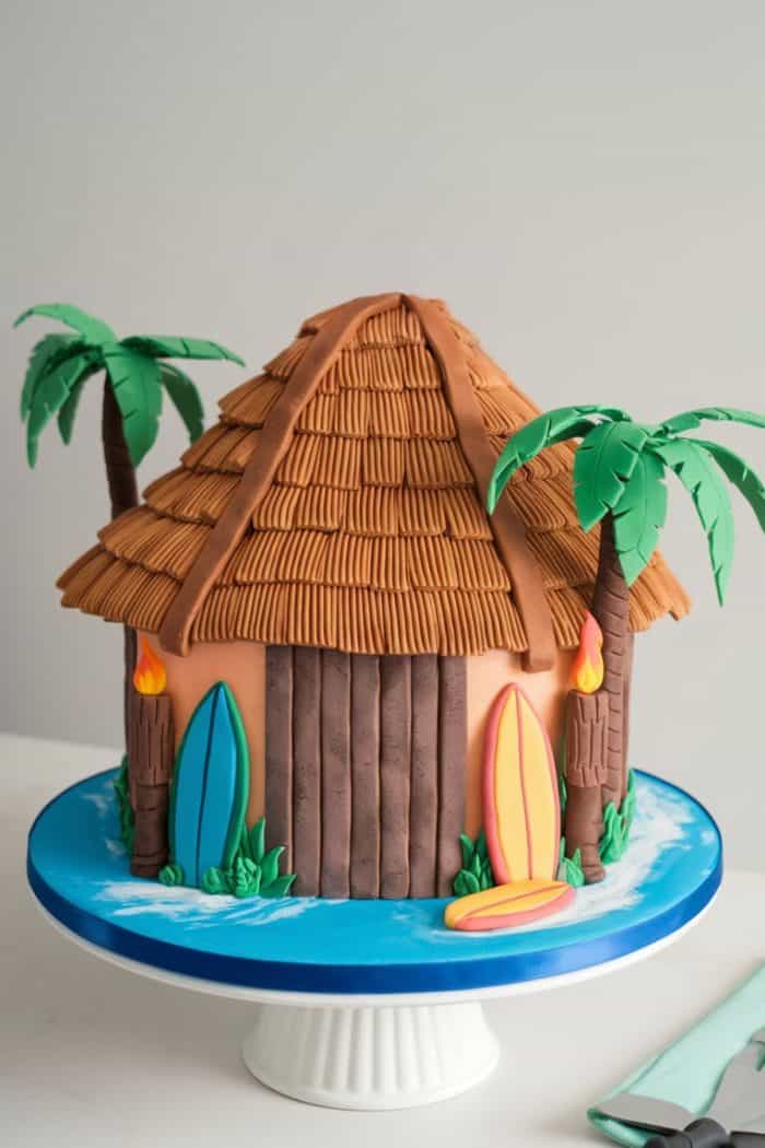 A cake designed to look like a tropical hut with a thatched roof. It features fondant palm trees, surfboards, and a sandy icing base, all on a blue cake stand.