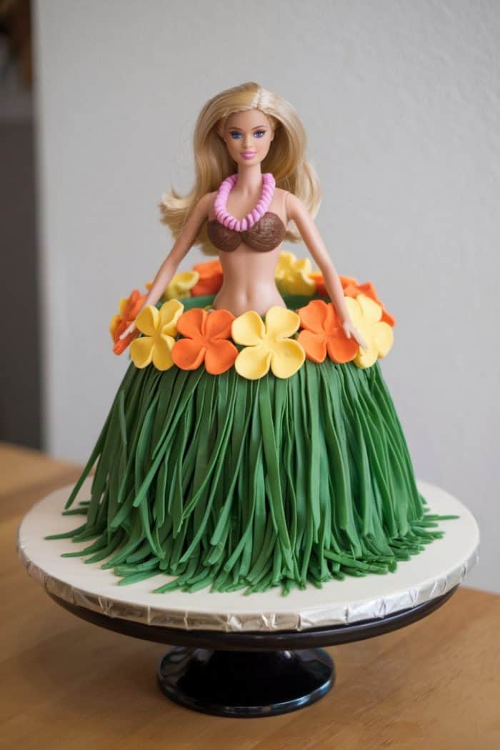 A cake designed with a doll on top, dressed in a hula outfit. The doll wears a brown top, green skirt made of frosting, and a lei of orange and yellow flowers. It's placed on a white cake stand.
