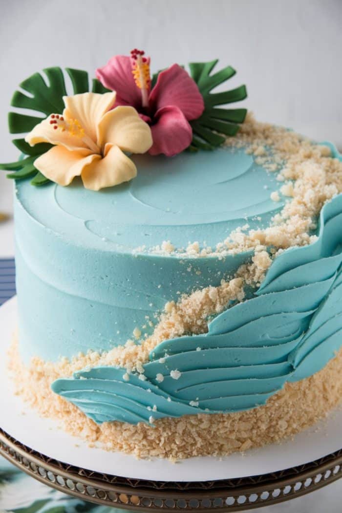 A blue frosted cake with wavy icing resembling ocean waves. Sand-like crumbs decorate the side, and the top features tropical leaves and hibiscus flowers in pink and yellow. The cake is displayed on a round metal stand.