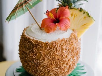 A cake designed to look like a coconut, topped with a red hibiscus flower, a pineapple slice, and a small decorative umbrella. It sits on a round pedestal with green leaves at the base.