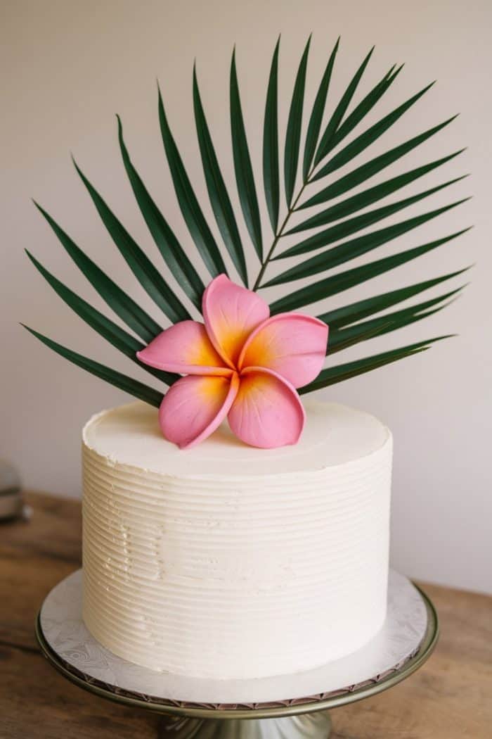 A white frosted cake on a stand, decorated with a large pink and yellow tropical flower and a green palm leaf on top, placed on a wooden surface with a soft background.