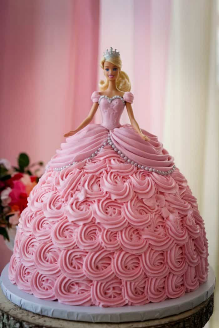 A cake designed as a princess doll in a pink dress with intricate frosting details. The doll wears a silver tiara and has blonde hair. The background is softly blurred in shades of pink and white.