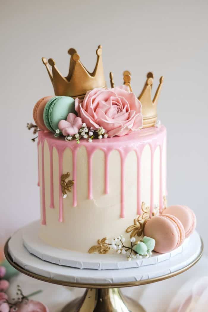 A decorated cake with pink icing drips, topped with a large pink rose, macarons in green and pink, and two golden crown toppers. Gold leaf accents and small flowers adorn the sides. The cake sits on a white and gold stand.
