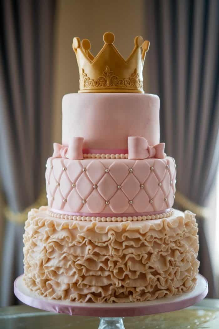 A three-tiered cake with a golden crown on top. The top layer is pink with a smooth finish, the middle layer is quilted with pearls and two pink bows, and the bottom layer features ruffled icing. The cake is displayed against draped curtains.