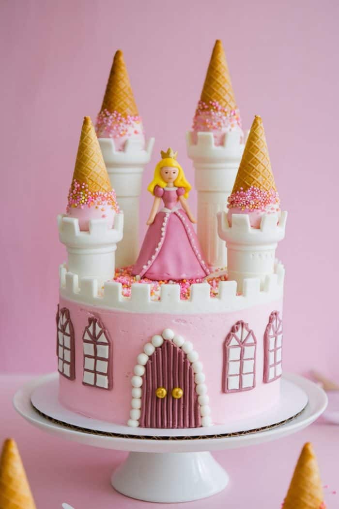 A pink princess-themed cake with castle turrets and ice cream cone spires topped with sprinkles. A fondant princess in a pink dress stands on top. The cake is on a white stand against a pink background.