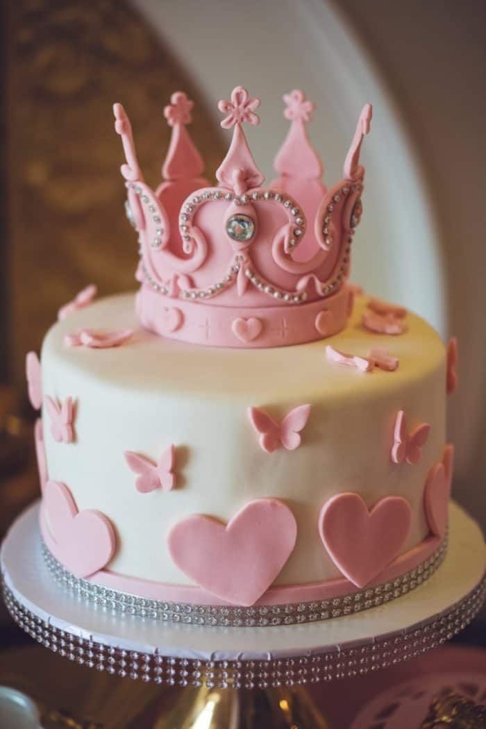 A white cake adorned with pink fondant decorations, including hearts and butterflies. The cake is topped with a pink crown decorated with small jewels. A shimmering band encircles the base of the cake.