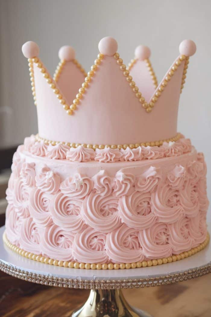 A pink cake with intricate, swirled frosting, topped with a large, pink crown. The crown is decorated with gold beads along its edges. The cake sits on a reflective silver cake stand.