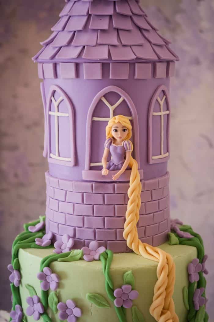 A cake designed like a tower with a purple and green base. A figure resembling a fairy-tale character with long braided blonde hair leans out of a window. Purple flowers decorate the cake, enhancing the whimsical theme.