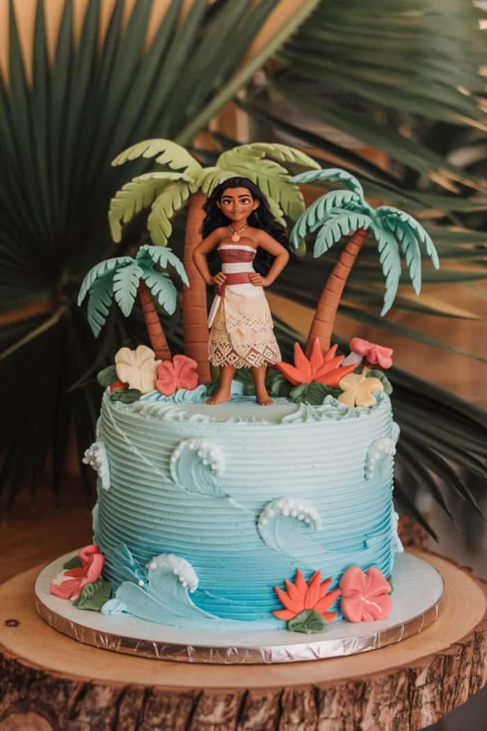 A blue cake decorated with ocean wave patterns features a topper of a girl in a patterned dress holding an oar, standing between two palm trees. Colorful flowers adorn the cake, with large green leaves in the background.