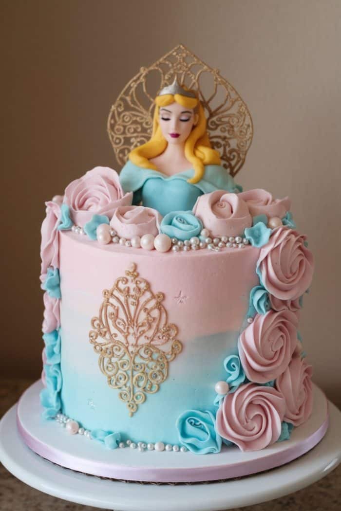 A cake with pink and blue frosting features a princess figure on top. Decorated with pink and blue roses, gold ornate accents, and white pearls. The princess has long blonde hair and a golden crown.