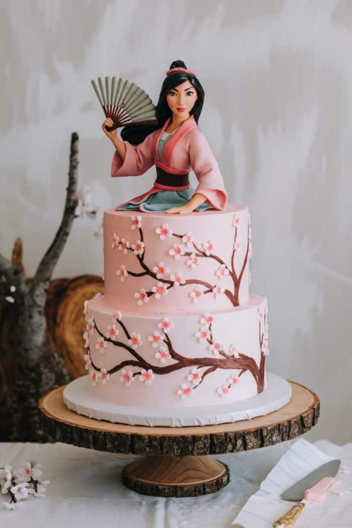 A two-tiered cake with pink icing and cherry blossom designs. On top, a figure of an animated character in traditional attire holds a fan. The cake is displayed on a wooden stand.
