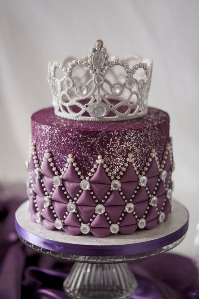 Elegant purple cake with a quilted design and pearl accents. The top is covered in glittery purple, crowned with an ornate silver tiara. Displayed on a glass cake stand with a draped purple fabric in the background.