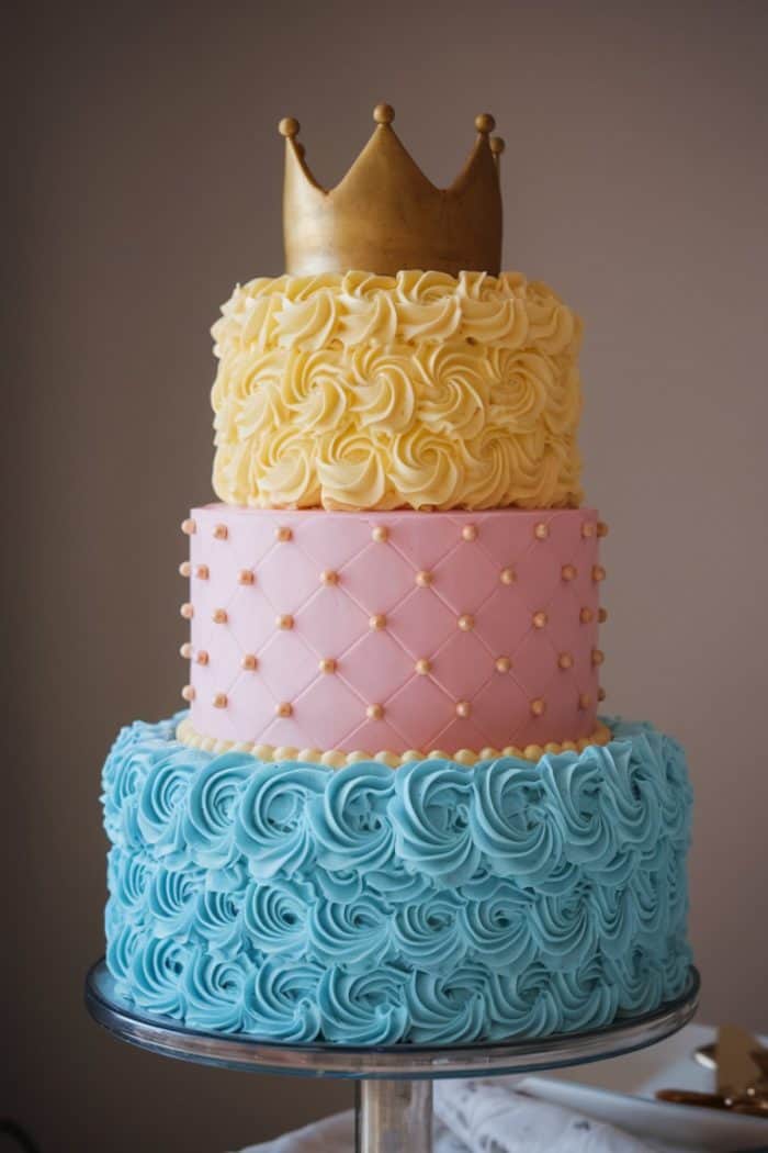 A three-tier cake decorated with intricate icing patterns. The bottom tier is blue with swirls, the middle is pink with a quilted pattern and gold dots, and the top is yellow with rosettes. A gold crown sits atop the cake.
